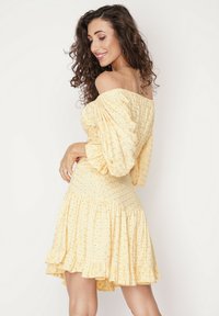 Żółta sukienka z dekoltem off-the-shoulder, teksturowany materiał z małymi wzorami kwiatowymi i falbaną na dolnej krawędzi. Krótka, dopasowana sylwetka.
