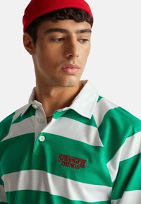 Zielono-biała koszulka polo w paski z kołnierzykiem; na przodzie czerwony haft "STRANGER THINGS". Gładka tkanina; guziki na plisie.