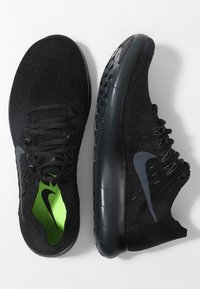 Chaussures de sport noires avec une tige en tricot texturé, des lacets noirs et un intérieur vert. Elles disposent d'une semelle foncée et lisse avec une traction à motifs.