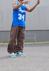 Canottiera da basket blu con lettering bianco, pantaloni cargo marroni e sneakers turchesi con dettagli bianchi, su uno sfondo grigio.