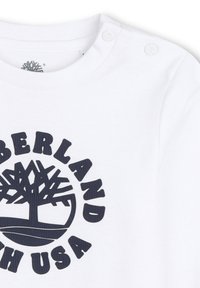 Witte katoenen lange mouwen shirt met een marineblauw rond boompje logo en de tekst "Timberland" en drukknopen op de schouder.