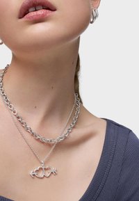 La joyería de plata presenta un diseño en capas con un collar de cadena gruesa y un colgante de dos corazones con una flecha. El tono de piel es neutro.