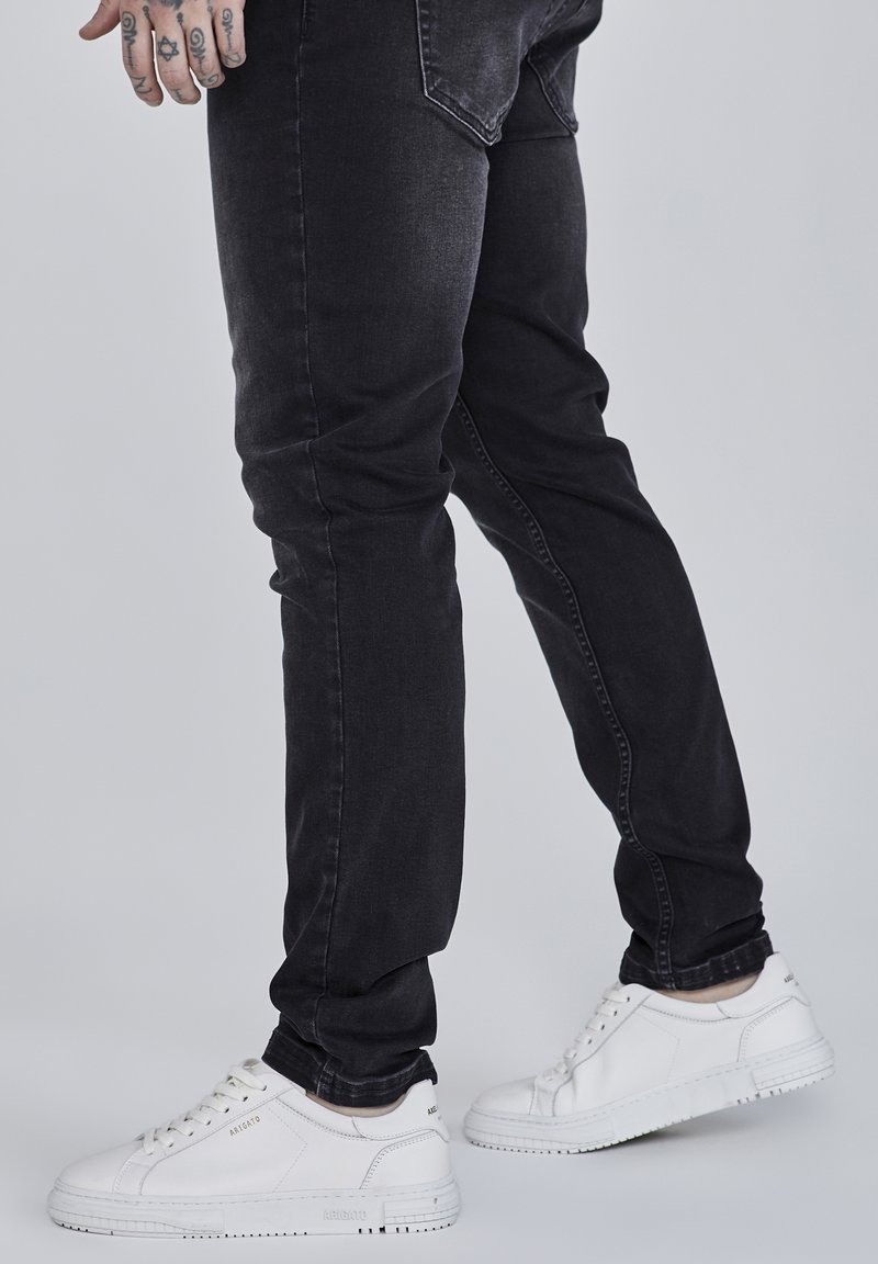 Homme portant un jean noir et des baskets blanches, montrant une main gauche tatouée reposant le long du corps sur un fond clair.
