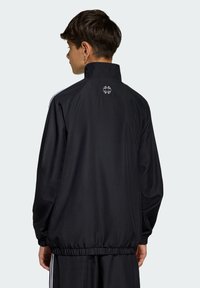 Chaqueta deportiva negra con cuello alto, cintura ajustable y rayas blancas laterales. Detalle de logo en la parte trasera, textura de tela suave.
