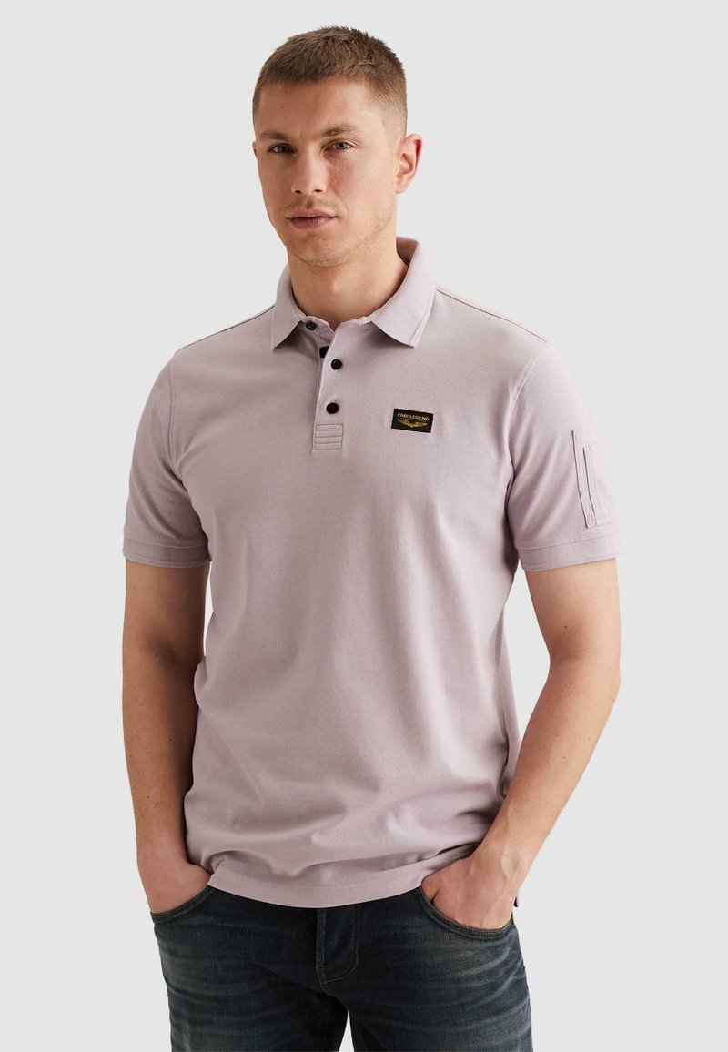 Jeune homme aux cheveux courts portant un polo violet clair avec des boutons noirs et un jean, mains dans les poches, fond neutre.