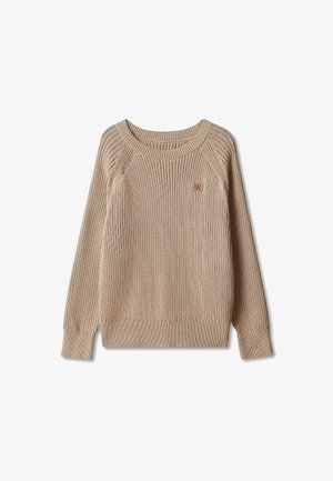 Lyst barn med krøllet hår iført beige striktrøje, sorte bukser og hvide sneakers, står med hænderne i lommerne på grå baggrund.