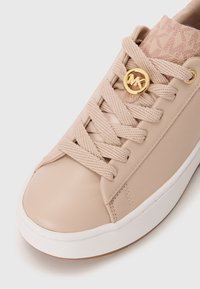 Baskets beiges avec des lacets épais, une semelle blanche et un pendentif circulaire doré avec le logo "MK" sur les lacets près de la languette.