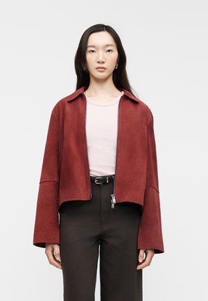 Γυναίκα που φοράει κόκκινο φούτερ με φερμουάρ και φαρδιά μανίκια, ανοιχτό ροζ μπλούζα, μαύρο παντελόνι και μαύρη ζώνη, στέκεται μπροστά σε μονόχρωμο φόντο.