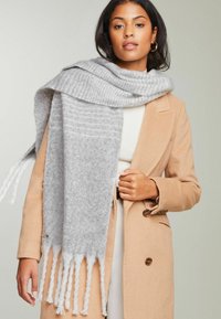Écharpe tissée grise avec une texture douce, présentant un motif et des franges blanches aux extrémités, posée sur un manteau beige.