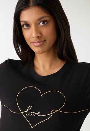 Młoda kobieta o długich ciemnych włosach, ubrana w czarną koszulę z konturem złotego serca i napisem „love” pisanym kursywą wewnątrz serca.