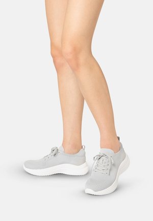 Ben iført lysegrå strikkede sneakers med hvide såler og snørebånd, stående på en hvid overflade mod en neutral baggrund.