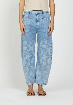 Woman wearing wide-leg blue jeans with large front pockets and faded floral patterns, paired with beige slide sandals.