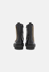 Botas de tobillo de cuero negro con suela gruesa, cremallera lateral y acento de cadena dorada a lo largo de la costura exterior. Textura suave y diseño elegante.