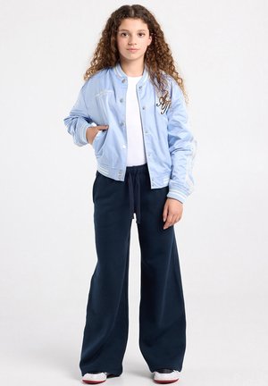 Jonge persoon met krullend haar, gekleed in een lichtblauwe bomberjack, wit shirt, wijde marineblauwe broek en witte sneakers, poserend met één hand in de zak.