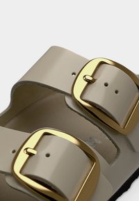 Κοντινό πλάνο μπεζ λουριών σανδαλιού με μεγάλες γυαλιστερές αγκράφες σε χρυσό τόνο και τρύπες ρύθμισης σκαμμένες σε λευκό φόντο.