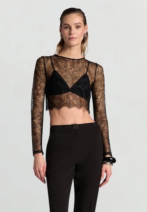 Top corto in pizzo nero con maniche lunghe trasparenti, sovrapposto a un reggiseno triangolare nero. Taglio aderente con orlo smerlato. Abbinato a pantaloni neri su misura.