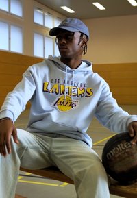 Svijetloplava dukserica s grafikom "Los Angeles Lakers", svijetle traperice, sivi kapu, drži crnu košarkašku loptu u teretani.