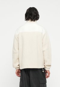 Veste en polaire avec un corps texturé crème et un col blanc. Dotée d'un cordon de serrage à l'ourlet, de manches longues et d'une coupe décontractée.