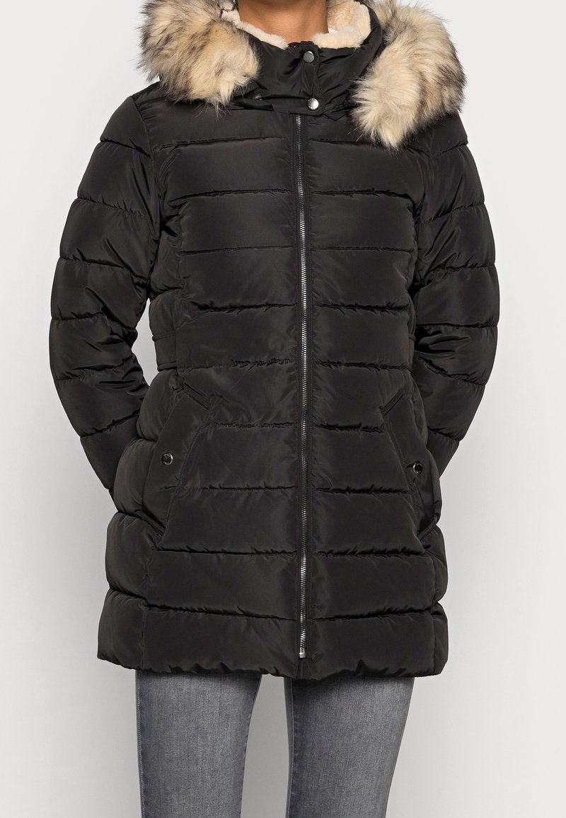 Femme portant un manteau d'hiver matelassé noir avec une fermeture éclair à l'avant, des poches latérales, et une capuche bordée de fourrure beige et noire.