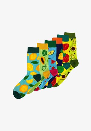 Jonge vrouw lacht, draagt een zwarte sportbeha, een blauwe geplooide spijkerrok en groene sokken met gele en oranje fruitpatronen.