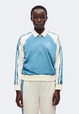 Personne portant une veste de sport zippée bleu clair et blanc avec un pantalon de survêtement blanc assorti avec des rayures bleues.