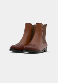 Brown leather ankle boots with brogue detailing, elastic side panels, and a rounded toe. Flat black rubber soles provide traction.