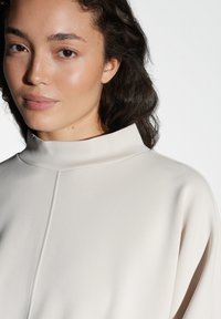 Top beige chiaro con un collo alto e una texture liscia e morbida. Presenta un sottile dettaglio di cucitura lungo la parte anteriore. Drappeggio elegante.