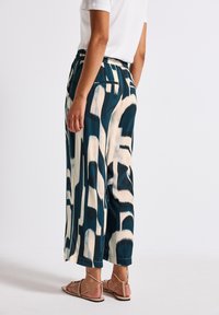 Weite Hosen in türkis und cremigem, abstraktem Muster, mit elastischem Bund und Seitentaschen. Das Material hat eine glatte, weiche Textur.