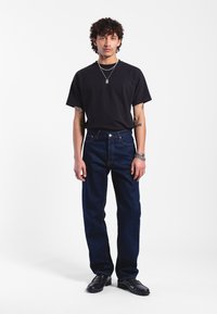 Black short-sleeve t-shirt, dark blue straight-leg jeans with contrast stitching, and black shoes. Model stands against a white background.
