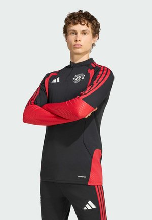 Młody mężczyzna ubrany w czarno-czerwony sportowy strój Manchester United Adidas, z założonymi ramionami, stojący na jednolitym jasnym tle.