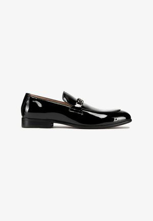 Zwarte gepatenteerde leren loafers met een gladde, glanzende afwerking, voorzien van een eenvoudige band en decoratieve hardware op de bovenkant. Lage, platte hak.