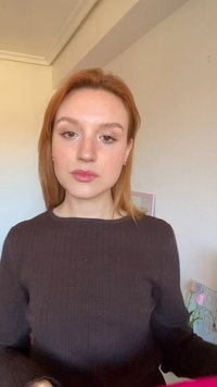 Jeune femme aux cheveux raides roux portant un pull marron, debout à l'intérieur près d'un mur avec des fleurs de tulipes et un tableau encadré.