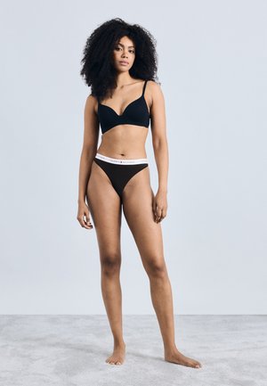 Donna con capelli ricci che indossa un reggiseno nero e biancheria intima Tommy Hilfiger nera, in piedi a piedi nudi su una superficie grigio chiaro contro uno sfondo semplice.