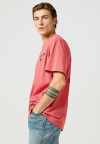 Camiseta de manga corta en algodón coral con cuello redondo. Presenta un logo negro en el frontal y detalles de texto adicionales en la parte posterior.