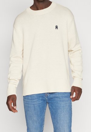 Man wearing cream knit sweater with ribbed cuffs and hem, paired with blue jeans, standing against a plain light background.