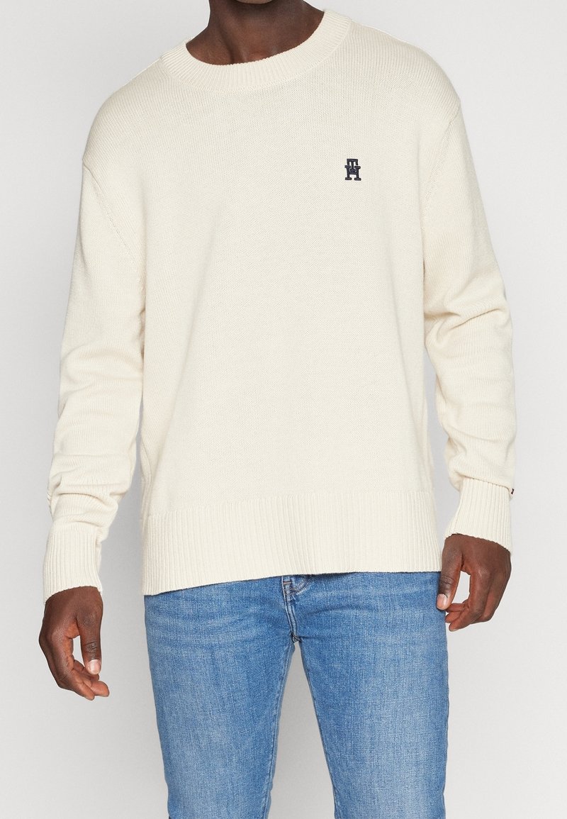 Homme portant un pull en tricot crème avec poignets et ourlet côtelés, associé à un jean bleu, debout devant un fond clair uni.