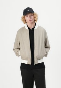 Beige bomberjakke med hvid ribkant ved kraven og ærmerne, med lynlås foran og sidelommer. Bæres over en sort tekstureret skjorte og bukser.