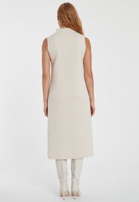 Ärmelloses gesteppte Kleid in Creme. Merkmale einen hohen Halsausschnitt, knielangen Schnitt und ein strukturiertes Muster. Kombination mit kniehohen weißen Stiefeln.