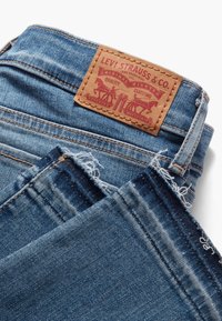 Jeans de mezclilla en un lavado azul medio con un dobladillo desgastado, que cuentan con una etiqueta de cintura de cuero marrón con diseño en relieve y detalles en rojo.