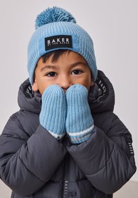 Mlad otrok, ki nosi svetlo modro pleteno kapo z cofkom in rokavice, ter temno puhovko, pokriva usta z rokami.