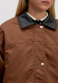 Bruine jas met een zwarte faux leren kraag, voorzien van drukknoopsluitingen en een gladde textuur. Het ontwerp heeft zichtbare stiksels en twee borstzakken.