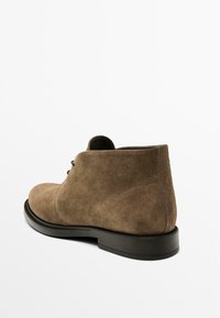 Stivali alla caviglia in suede marrone con una finitura liscia, punta tonda e suola in gomma nera. Presentano lacci e un sottile dettaglio di cucitura sul retro.