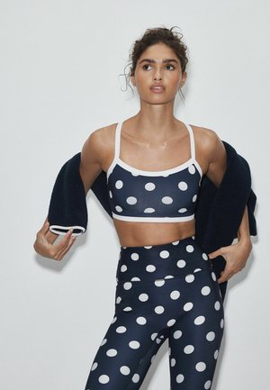 Femme portant un soutien-gorge de sport bleu marine à pois blancs et des leggins, tenant une veste foncée sur ses épaules devant un fond uni.