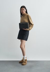 Zwarte leren handtas met gouden ritssluiting, doorgestikt ontwerp, model draagt een beige coltrui, donkere spijkerrok en bruine schoenen met bontvoering.