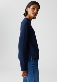 Femme aux cheveux courts et foncés portant un haut en marinière navy à manches longues texturé et un jean bleu, se tenant de profil contre un fond gris clair.