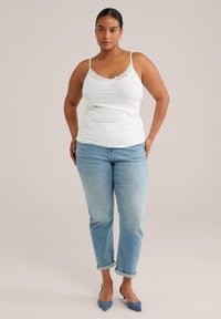 Mujer de pie con las manos en los bolsillos, vistiendo una camiseta de tirantes blanca con encaje, pantalones vaqueros claros arremangados y tacones azules de punta en un fondo liso.