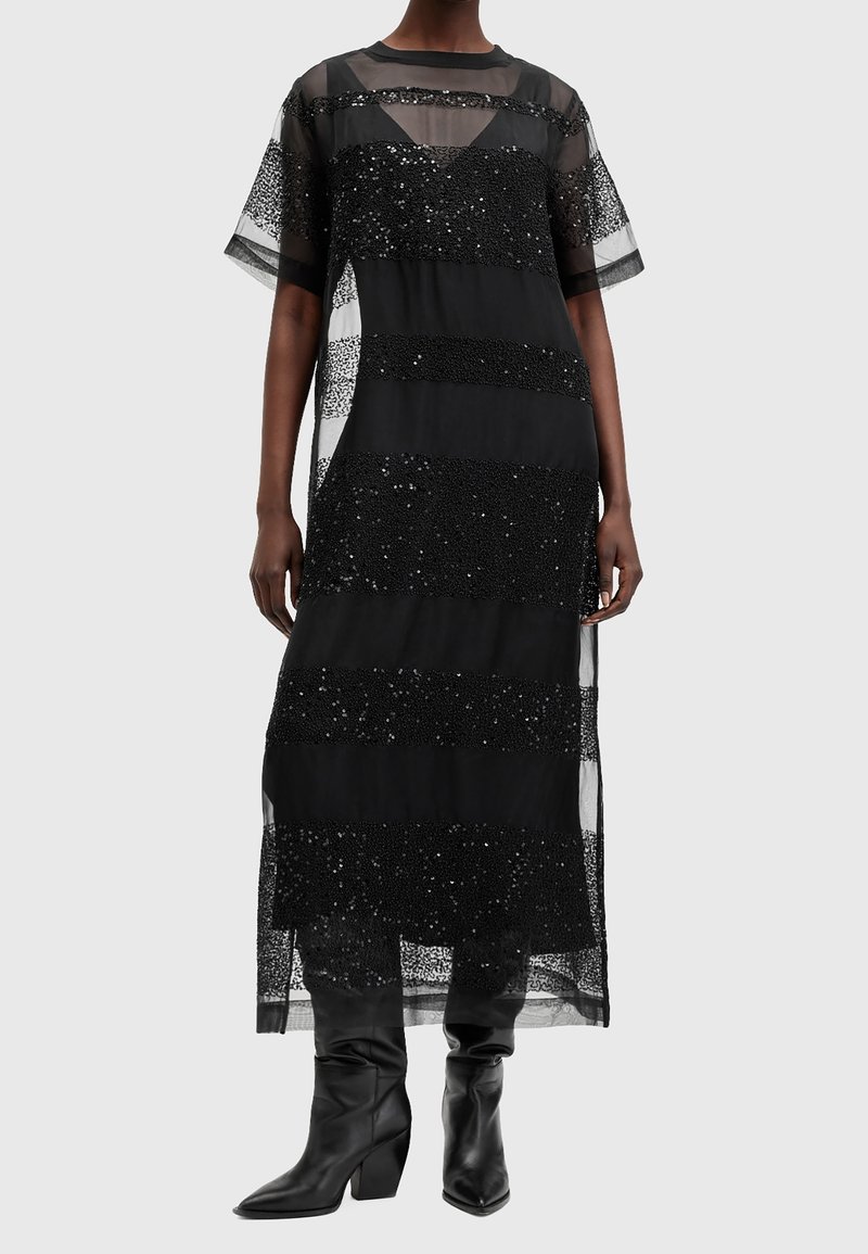 Robe en maille noire, longue, avec des rayures de sequins horizontales, des manches courtes et un design superposé. Associée à des bottes noires à talons montant jusqu'aux genoux.