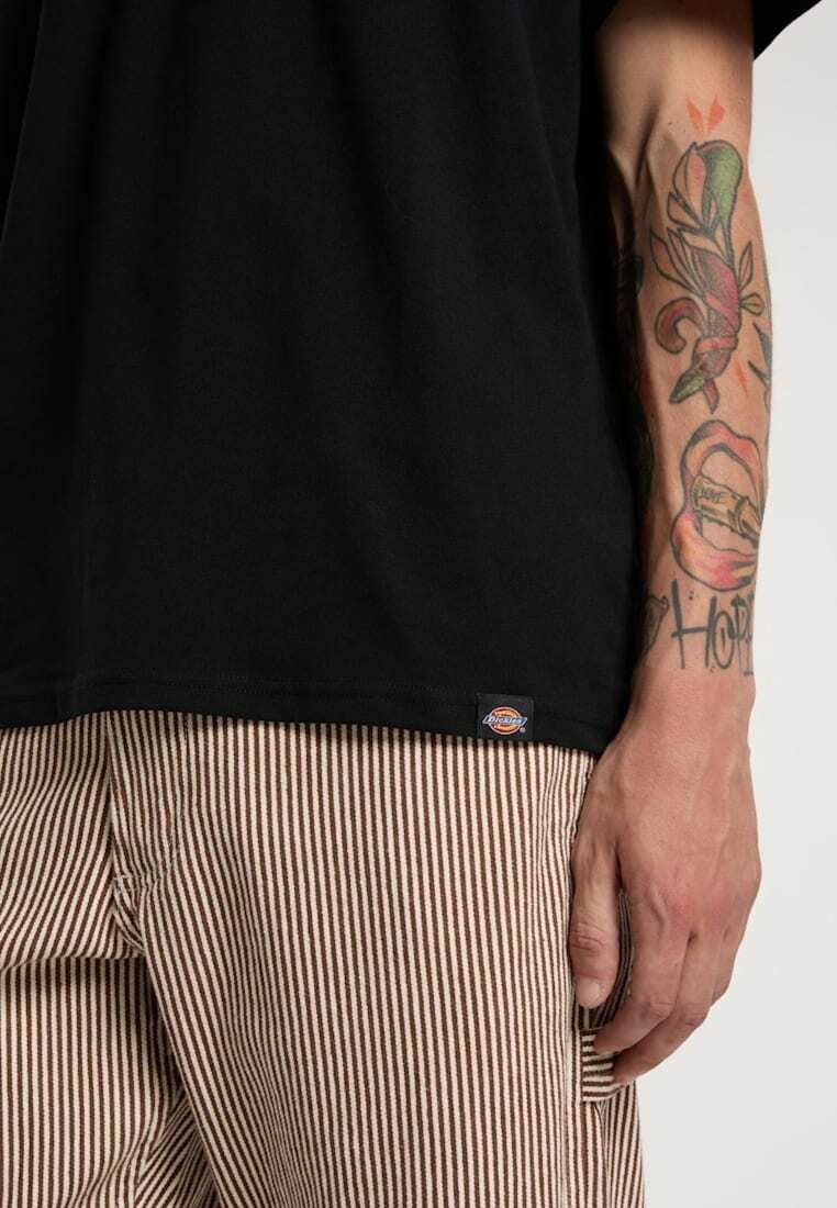 Mandens højre arm med farverige tatoveringer, iført en sort skjorte og beige og hvide bukser med lodrette striber.
