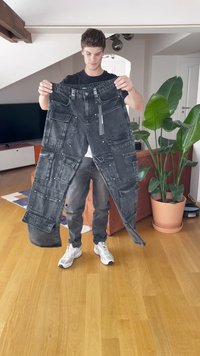 Black denim pants with multiple pockets, textured detailing, and a slightly distressed finish, held by a person in a bright room.