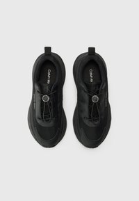Chaussures de sport noires avec une tige texturée, des lacets élastiques et une semelle en caoutchouc. Présente des logos sur la semelle intérieure et sur le côté. Design simple et moderne.